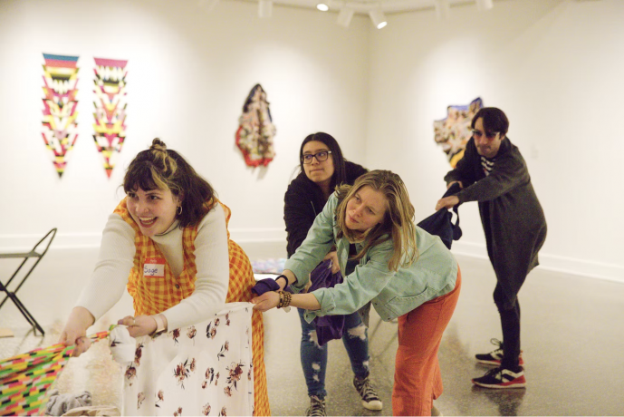 4 individuals perform in an art gallery space. They each hold on to a multicolored fabric rope and are tugging it and smiling. 